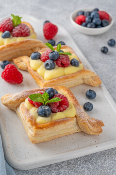 Danish puff pastry cakes with custard cream and fresh berries, sprinkled with powdered sugar on a white ceramic board on a concrete background.