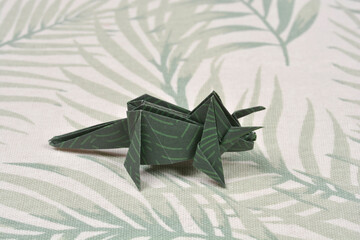 A handmade dark green origami triceratops with a light green leaf pattern stands on a textured fabric background featuring palm leaf prints © nahhan