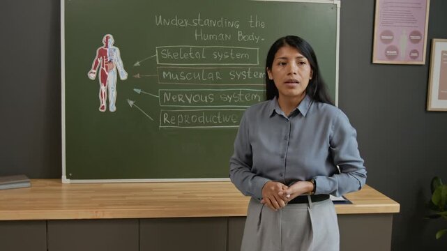 Medium full dolly-out shot of Hispanic female teacher drawing with chalk on board while taking about human body systems and reproductive health to class of teenage school students