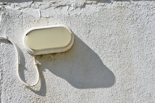 A weathered oval white bulkhead lamp is mounted on a rough textured white building facade with a visible power cable and a long sharp shadow