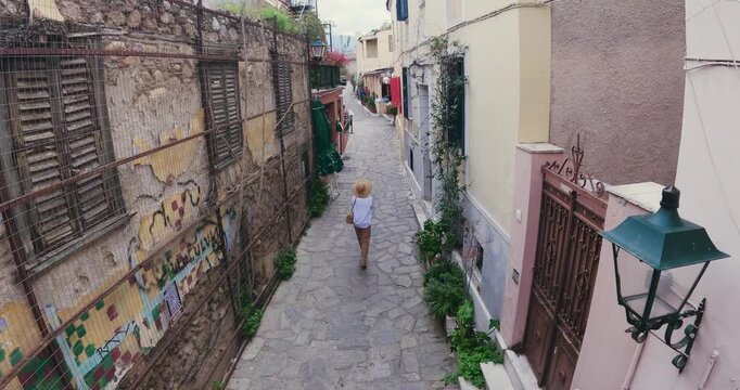 Athens, Greece - Mar 10, 2024: Tourist woman wearing hat walk on narrow street of Anafiotika neighborhood and Acropolis in the old town of Athens, Greece, aerial drone shot