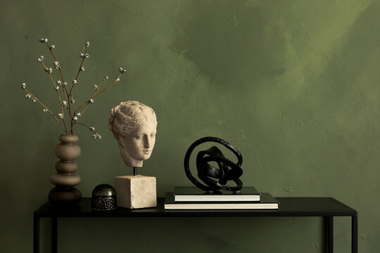 Stylish arrangement with modern decor, bust sculpture, willow branches, and decorative objects on a console table against a green wall