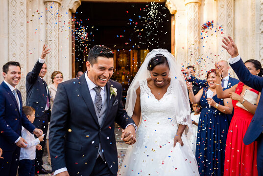Joyful interracial bride and groom laughing while walking through confetti outside a church. Happy diverse wedding couple leaving ceremony with guests. Celebration of marriage.