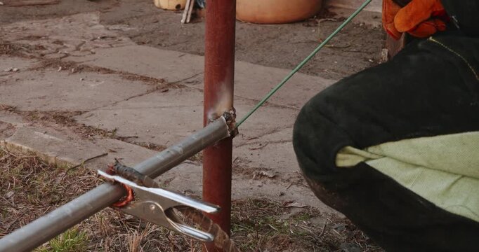 Industrial worker welding metal pipes for a fence using an electric arc machine, with bright sparks flying from the molten metal joint as the electrode melts and fuses the structure together.