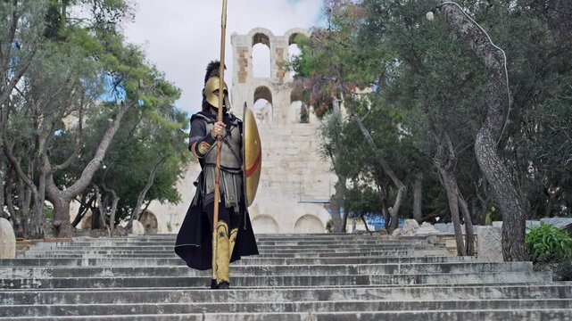 Confident hoplite spartan soldier wearing metal helmet and protecting with shield attacking with sharp spear with view of Acropolis Odeon of Herodes Atticus at background