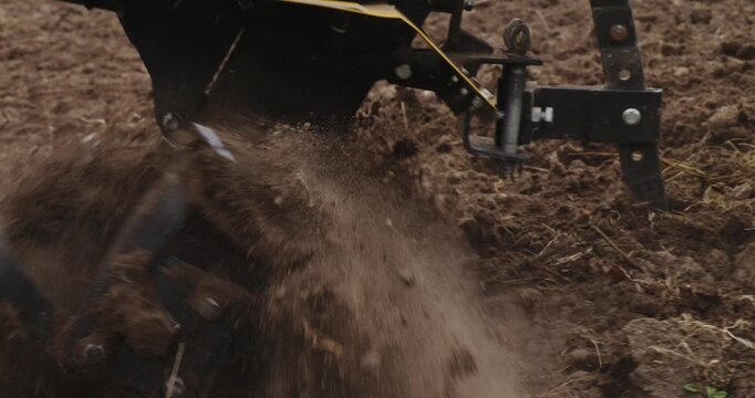 Close-up view of a power tiller's rotating blades digging into the earth, cultivating the soil in a field for spring planting and preparing the garden bed for a new crop.