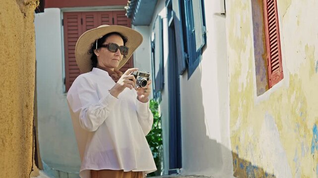 Fashion model stylish woman with hat and glasses stroll on narrow streets of Anafiotika under Acropolis in Athens, Greece taking a pictures with vintage retro camera