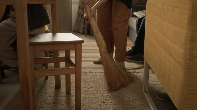 Close up low angle tilt up shot of unrecognizable woman sweeping floors with broom while her family reading in cramped but cozy apartment