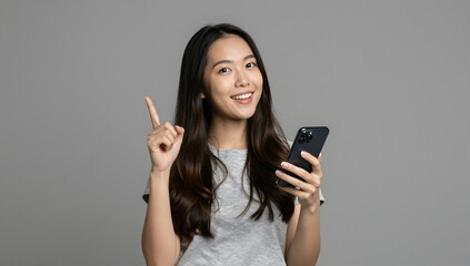 Fototapeta premium Asian Woman with Smartphone: A young Asian woman smiles confidently while holding a smartphone and pointing upwards with her index finger, conveying a sense of idea or direction.