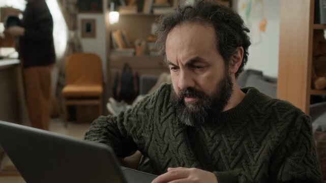 Stressed middle aged bearded man looking for job on his laptop while his wife washing dishes in kitchen in cramped, run-down apartment