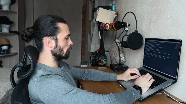 Recognizable Caucasian Young Man with Bun Hair Sitting at Wooden Desk and Typing on Laptop in Home Office Bedroom Studio Interior Sunny Day Daylight Eye Level Side View Profile Perspective Shot