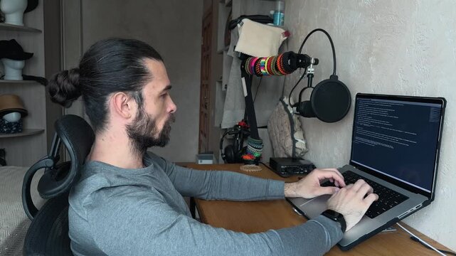 Recognizable Caucasian Young Man with Bun Hair Sitting at Wooden Desk Leaning Back with Hands Behind Head then Starting to Type on Laptop in Home Office Studio Side View Profile Eye Level Day Shot
