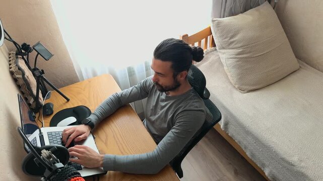 Recognizable Caucasian Young Man with Man Bun Hair Leaning Back in Mesh Chair with Hands Behind Head then Starting to Work and Type on Laptop in Home Office with Shifting Natural Daylight High Angle