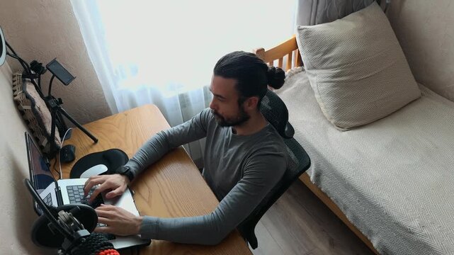 Recognizable Caucasian Young Man with Man Bun Typing on Laptop Stretching Tired Arms and Fingers Leaning Back in Mesh Chair Swaying and Thinking in Home Office High Angle Tilted Daylight Shot