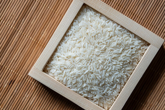 Raw white long grain rice in a square wooden box on a bamboo mat background. Healthy dry food staple ingredient for Asian cuisine