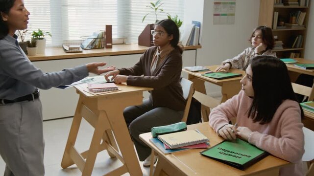 Full panning shot of young female teacher directing multiethnic teenage high school students to open textbooks on health education and perform test on understanding of new lesson material