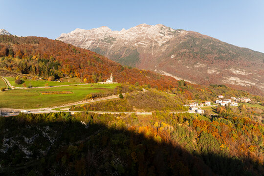 Autumn mountain landscape featuring San Tommaso church and Cavedago village, surrounded by vibrant fall forests, ideal for tourism and destination travel in Paganella, Trentino, Italy.