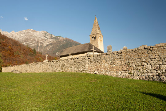 San Tommaso church, Cavedago, Paganella, Trentino, Italy, stands historically behind a stone wall with autumn mountains under a clear blue sky, ideal for tourism and nature travel, with copy space.