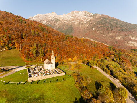 San Tommaso church and cemetery nestled in a vibrant autumn mountain landscape with a winding road and majestic peaks, a serene travel destination in Cavedago, Paganella, Trentino, Italy.