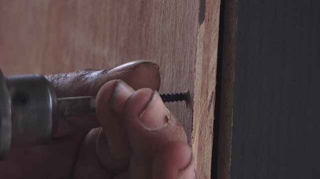 A detailed close-up shot showing a man&rsquo;s hands working on a DIY project. The right hand operates a cordless power drill to drive a screw into a piece of plywood, while the left hand stabilizes the scr