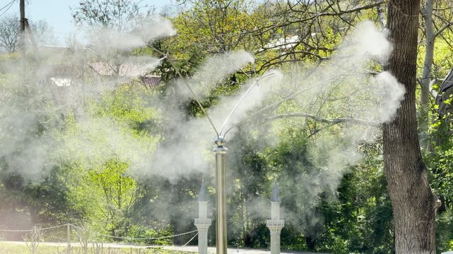 A metal misting system sprays fine water droplets into the warm garden air. The continuous spray creates a soft, soothing hiss. High-pitched bird songs echo through the surrounding green trees.