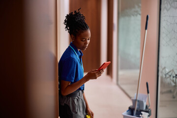 Black woman using cell phone while working as professional cleaner in office building.