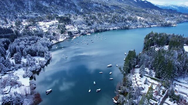 Static drone shot with a vertical camera tilt up, starting from a snowy bay filled with moored boats to reveal the vast Nahuel Huapi Lake and high snow-capped Andes mountains in Patagonia.