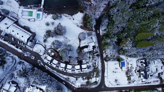 Drone moving forward with a vertical top-down camera view, traveling from a snow-covered area and forest toward the crystal clear turquoise waters of Nahuel Huapi Lake in the Andes.