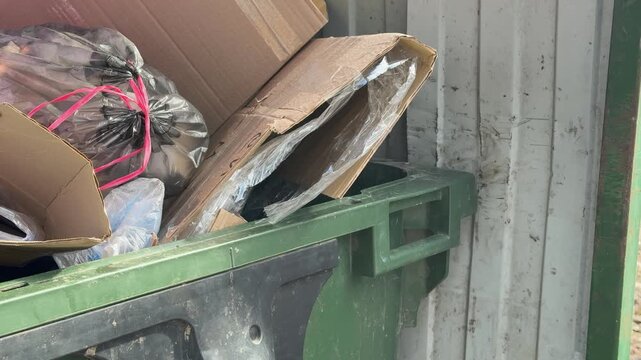 Close-up overflowing green dumpster loaded cardboard and construction debris including insulation, taped bags, crushed packaging