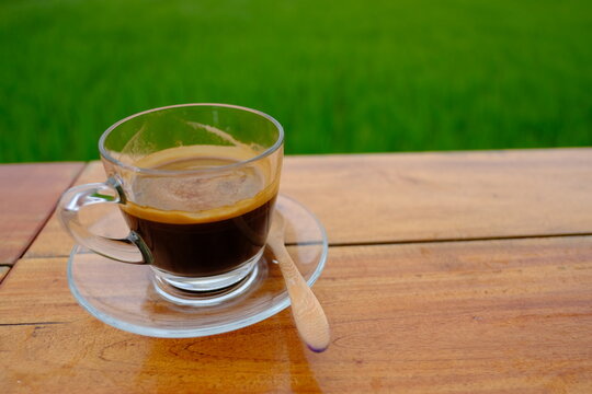 A cup of hot black coffee on a wooden table with beautiful green rice field and mountain background in the morning.
