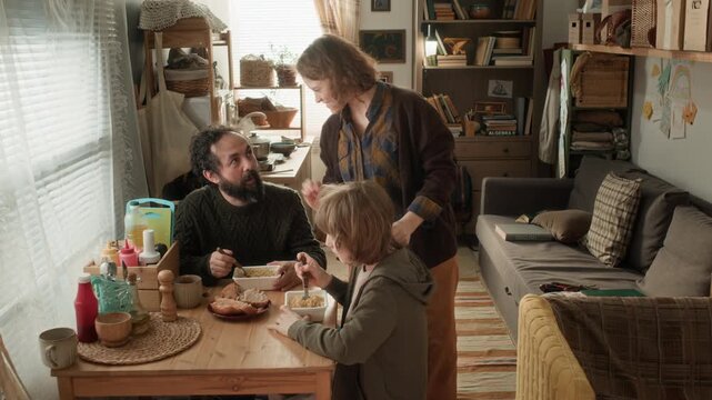 High angle shot of woman serving food to her family sitting at small wooden table in poor apartment