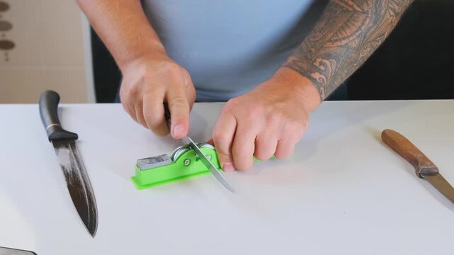 Man sharpening knife using whetstone on table, maintaining blade edge with manual sharpening technique, tool care and maintenance process at home, close up of hands and cutting tools