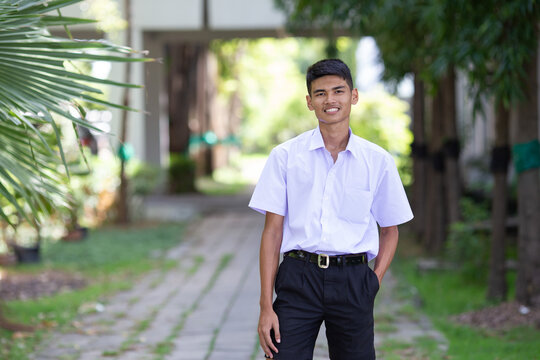 Thai boy student in official Thai school uniform (black shorts), ordinary appearance with slightly dark complexion, stand firmly on a walkway with one hand in his pocket, landscape orientation