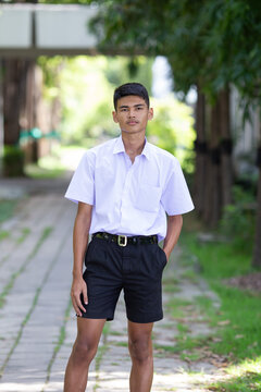 Thai boy student in official Thai school uniform (black shorts), ordinary appearance with slightly dark complexion, stand firmly on a walkway with one hand in his pocket, portrait orientation