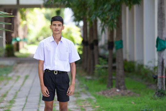 Thai boy student in official Thai school uniform (black shorts), ordinary appearance with slightly dark complexion, stand firmly on a walkway with arms at his sides, landscape orientation