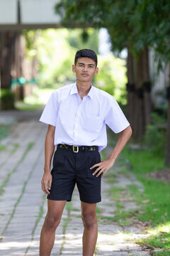 Thai boy student in official Thai school uniform (black shorts), ordinary appearance with slightly dark complexion, stand firmly on a walkway with one hand on his hip, portrait orientation