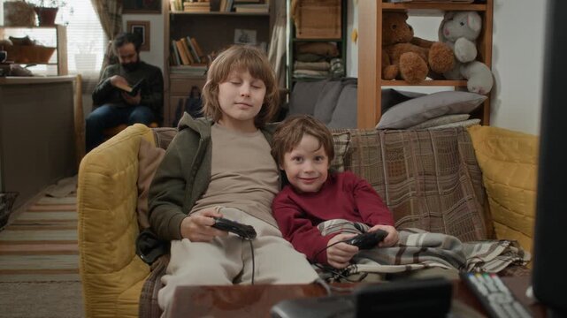 Low angle portrait shot of two brothers sitting on couch holding retro game controllers and smiing for camera in rundown cluttered apartment