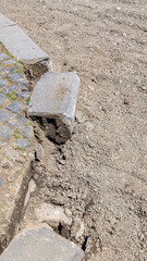 Fototapeta premium Road construction work showing a broken concrete curbstone being replaced, with excavated gravel, dirt, and stone pavement, symbolizing restoration and infrastructure development