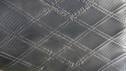 Fototapeta premium Dark perforated quilted fabric with diagonal stitches forming repeating diamond and rhombus motifs, showcasing textured, weathered surface with vintage, worn character