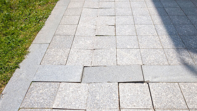 Damaged sidewalk paving causing a trip hazard, uneven gray granite blocks showing cracks and settling, requiring urgent repair and maintenance for pedestrian safety