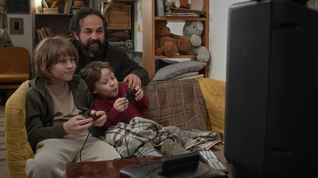 Father and his two sons enthusiastically playing video games on couch in front of big retro TV in poor apartment