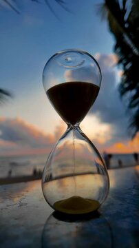 Hourglass on the beach at sunset with sand falling, symbolizing passing time, deadline, and life journey against a beautiful tropical sea background. 4K resolution, vertical.