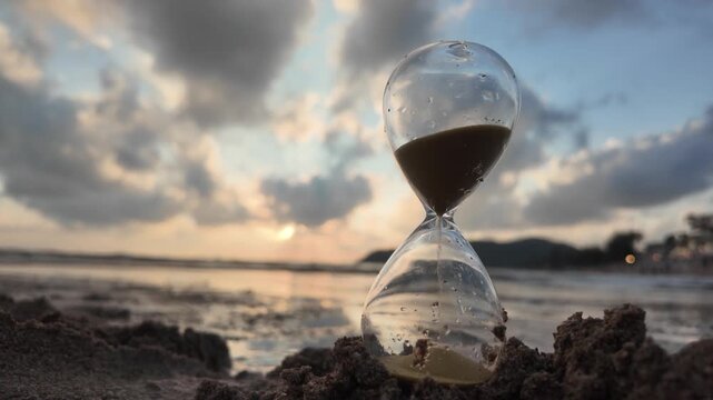 An hourglass on the beach with gentle waves flowing around it at sunset. Concept of passing time, fleeting moments, life transition, and time management. 4K slowmotion footage.