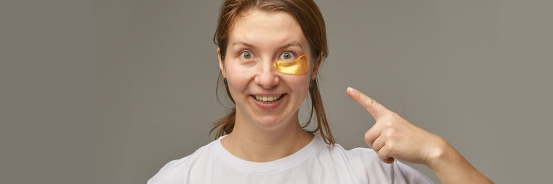 Woman wearing under eye patches pointing at face demonstrating skincare routine and cosmetic treatment isolated on neutral background