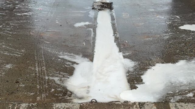 Foam wash over stained concrete floor, foam cascades from drain nozzle, thick white suds spread across textured pavement, wet