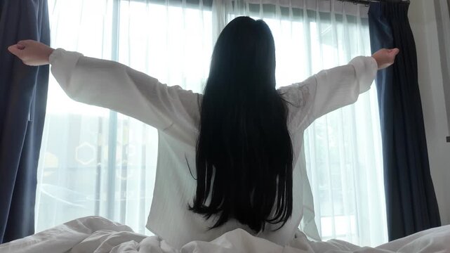 Slow motion of rear view of young woman wearing white pajamas, sitting on a bed and stretching relaxing in the morning. Concept of relaxation and healthy routine. 4K horizontal resolution