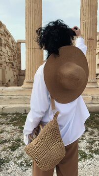 Young fashion woman visiting ancient Acropolis in Athens a Greek monument with stone columns, wearing white shirt, hat and stylish glasses