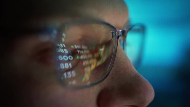 In dimly lit trading floor at night, focused trader wearing glasses watches his PC monitor, tracking real-time exchange rates price swings. Numbers flash across his lenses as he concentrates finance