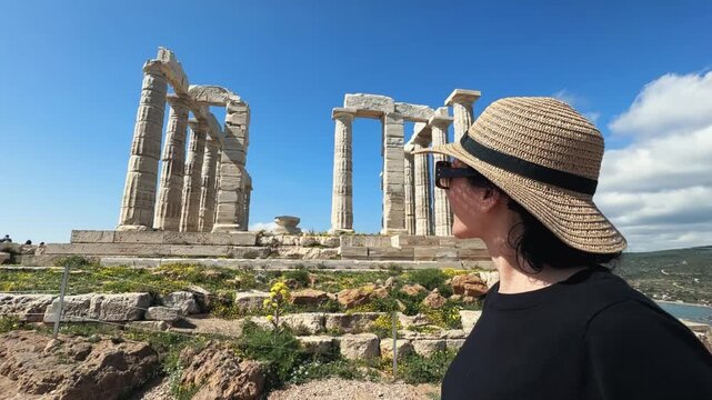 Stylish female tourist in black wearing hat and glasses enjoying sightseeing an ancient temple of Poseidon, Cape Sounion near Athens in Greece on summer sunny day