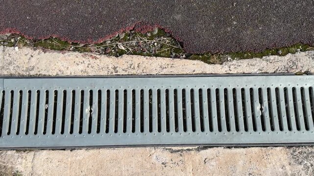 Galvanized drainage grate on asphalt, linear perforated metal slats running along track joint at stadium street closeup top down